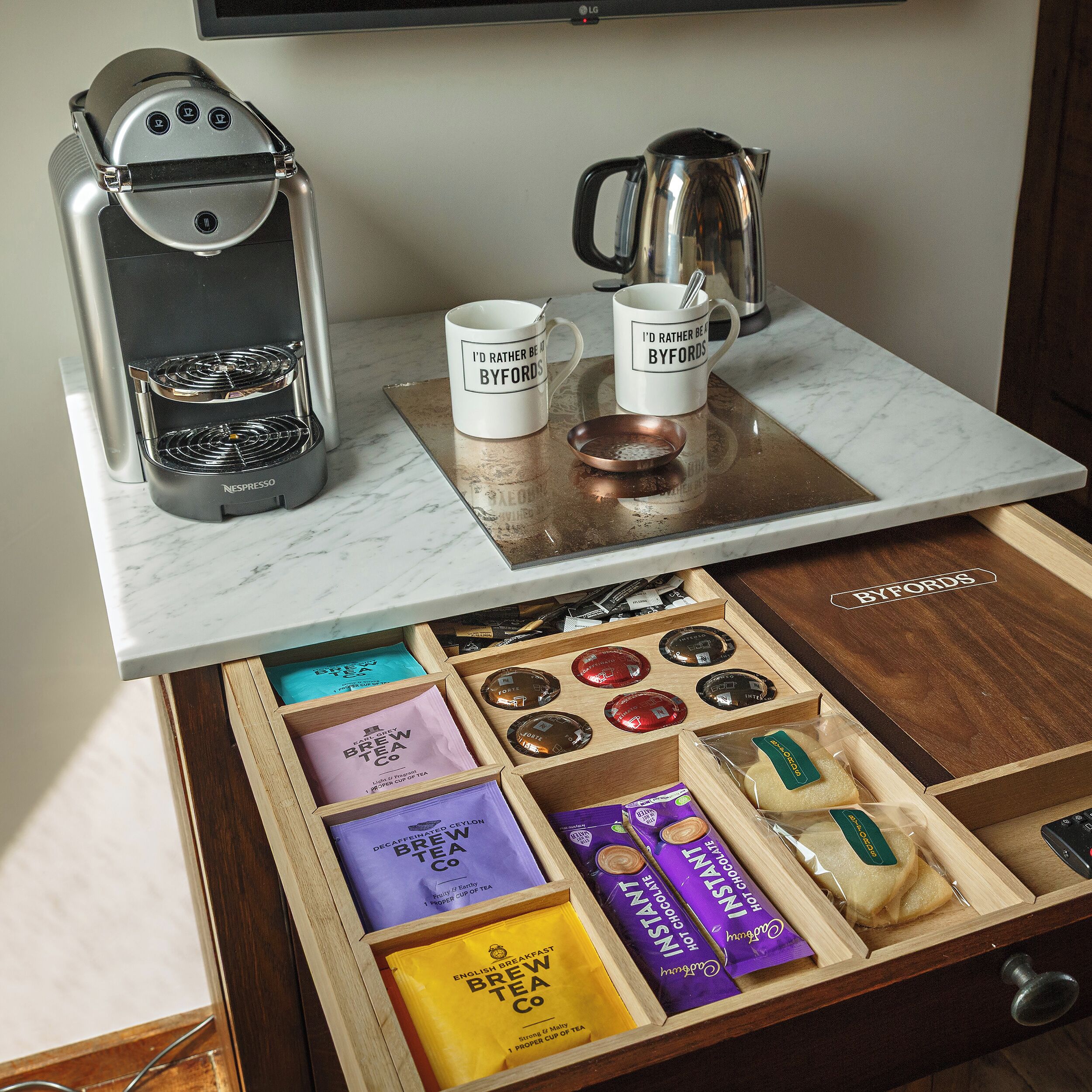 Una estación de café con una máquina Nespresso, dos tazas, un hervidor y una bandeja con diferentes productos de té y chocolate.