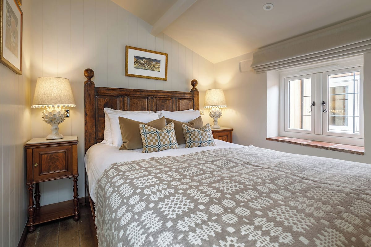 A bedroom with a large bed, two bedside tables with lamps, a window with curtains, and a framed picture on the wall.