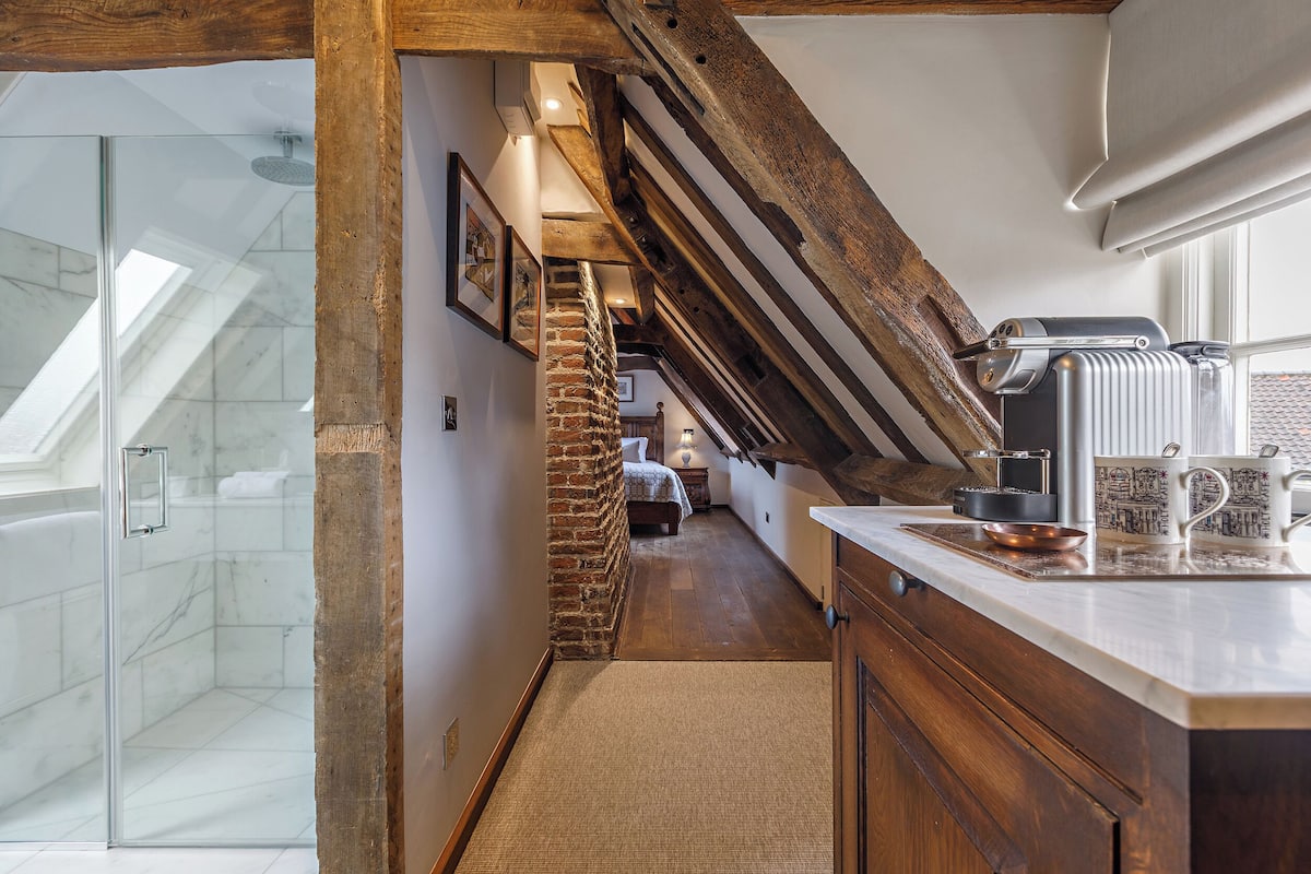 A narrow hallway with a glass shower, a brick column, wooden beams, a bed, and a kitchen area with a kettle and mug.