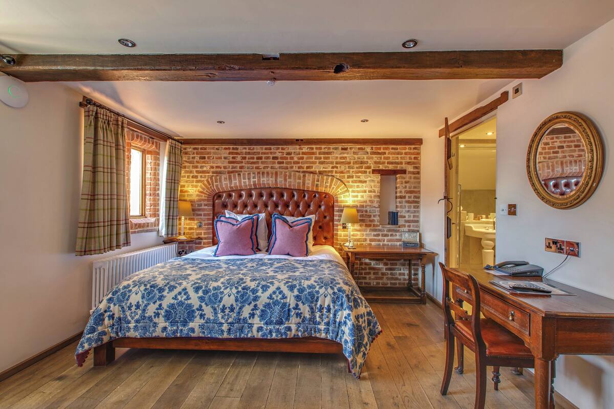A bedroom with a brick wall, a wooden headboard, a bed with a blue and white patterned bedspread, a wooden nightstand, a desk with a chair, and a mirror on the wall.