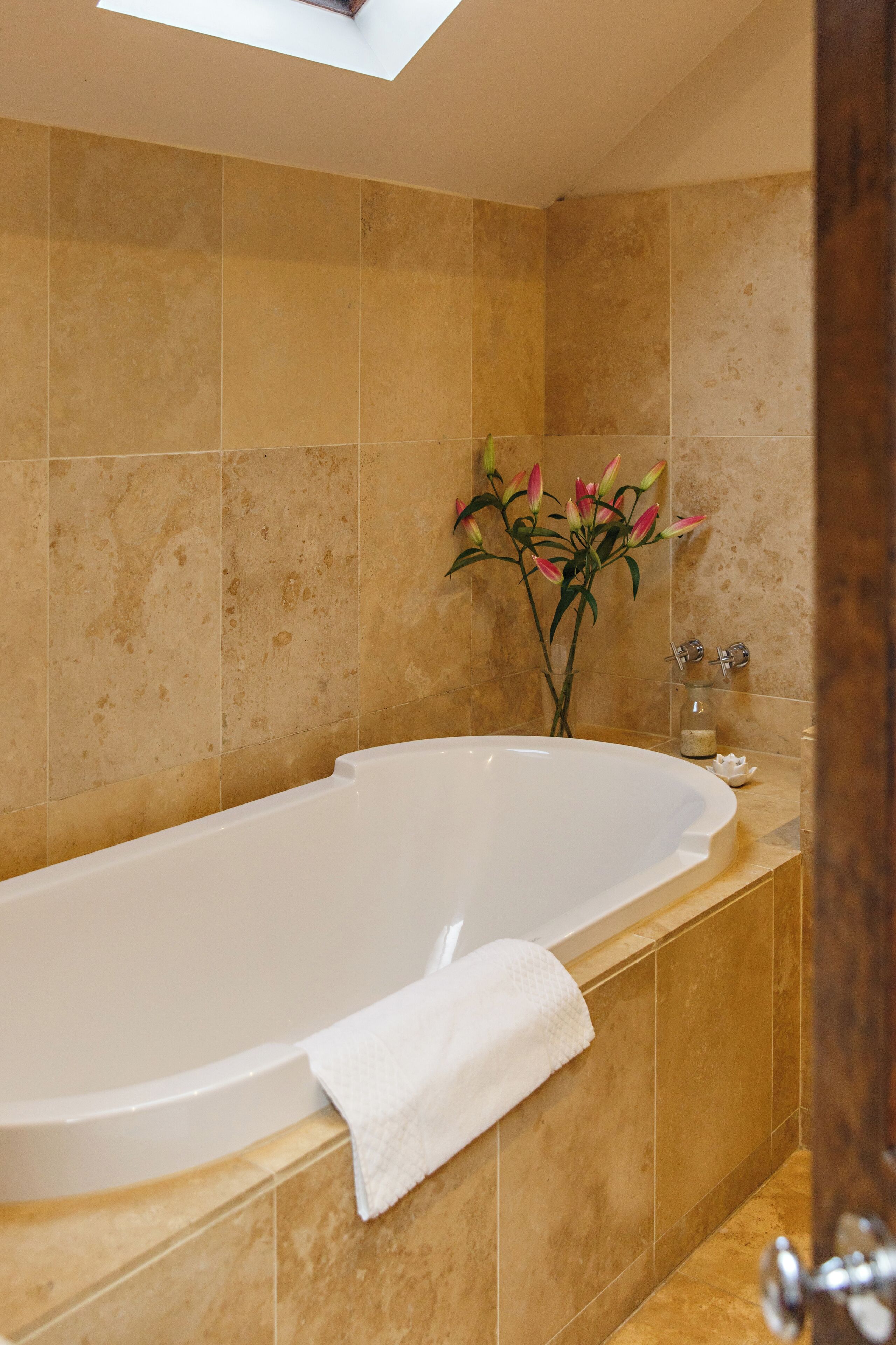 Un baño con una bañera grande, azulejos color beige, un florero con flores y una toalla sobre la bañera.