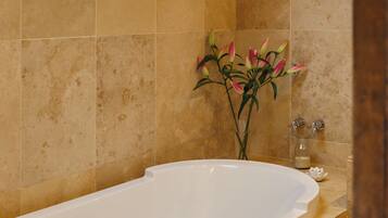 Un baño con una bañera grande, azulejos color beige, un florero con flores y una toalla sobre la bañera.