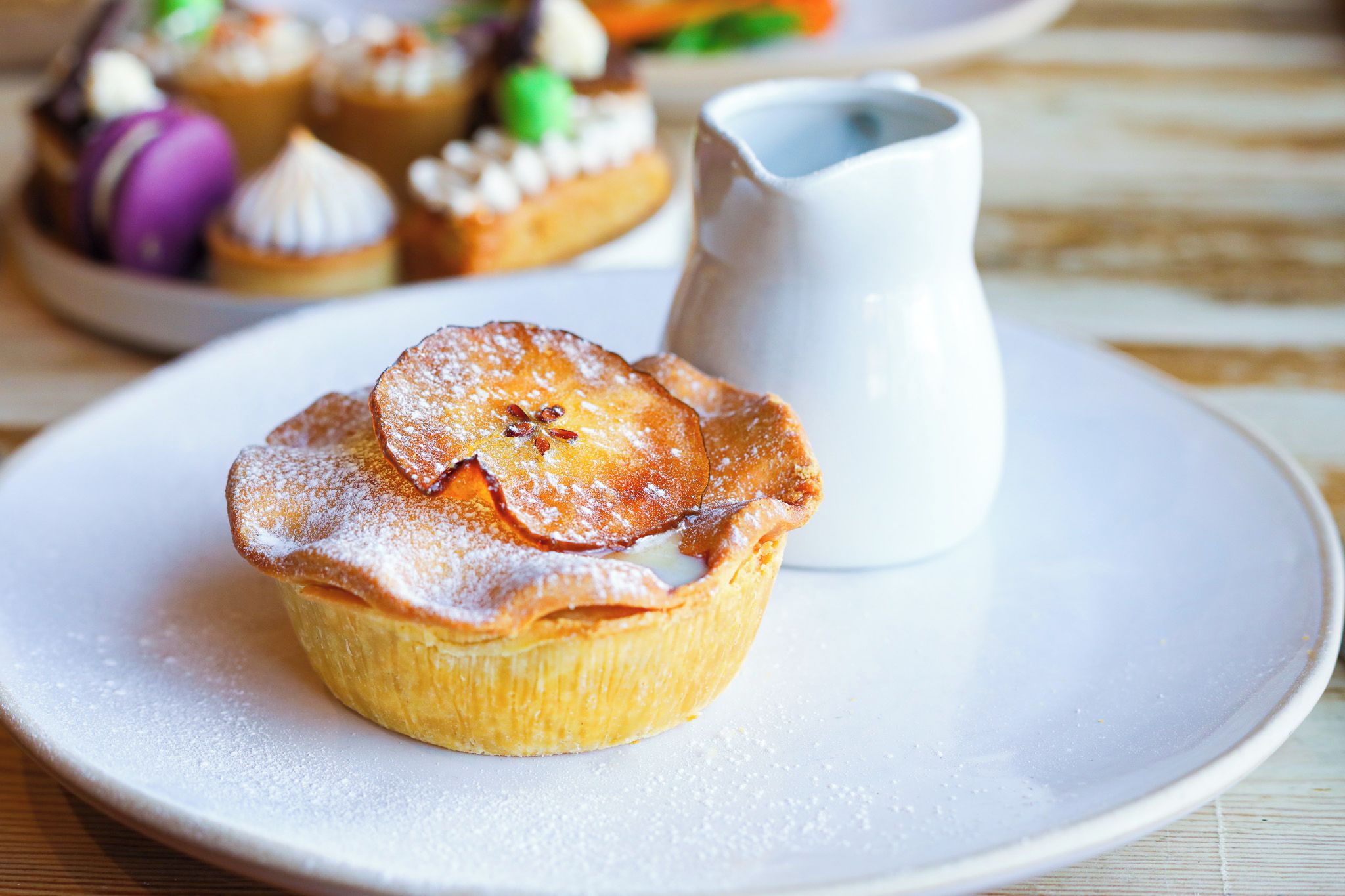 A dessert with a flaky crust and powdered sugar on a plate, accompanied by a small pitcher.