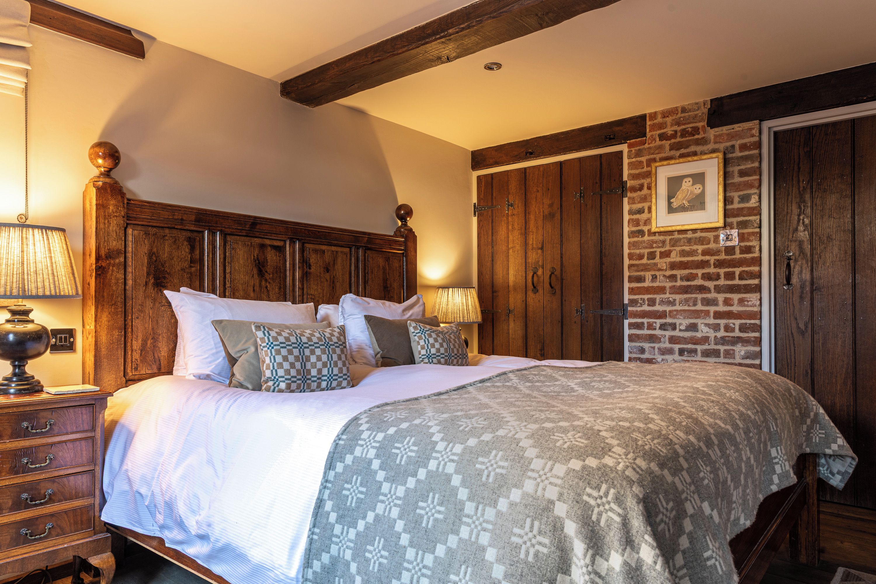 A bedroom with a large bed, wooden headboard, a nightstand with a lamp, a dresser, and a brick wall with a door.