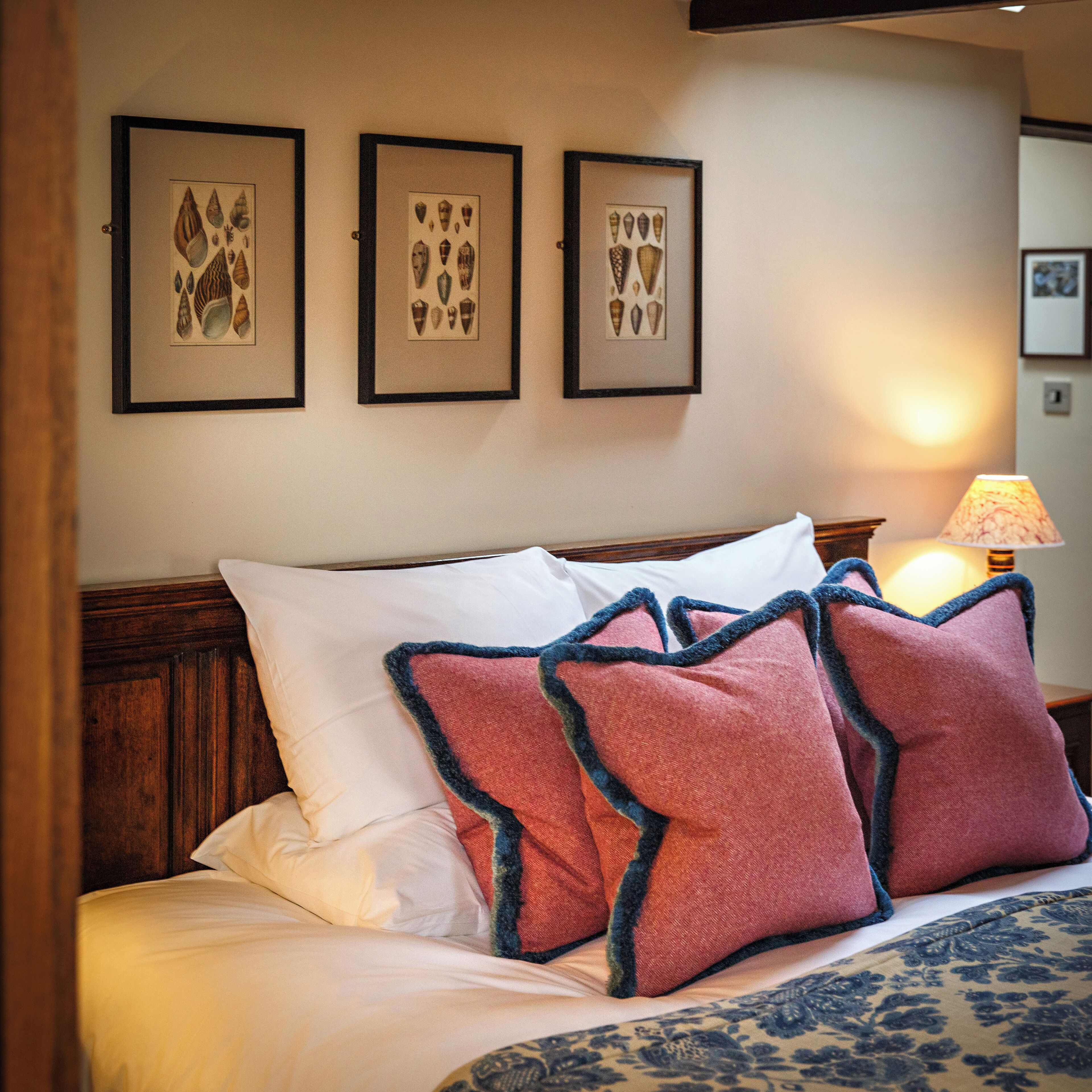 A neatly made bed with white and red pillows, a wooden headboard, and three framed botanical prints on the wall.