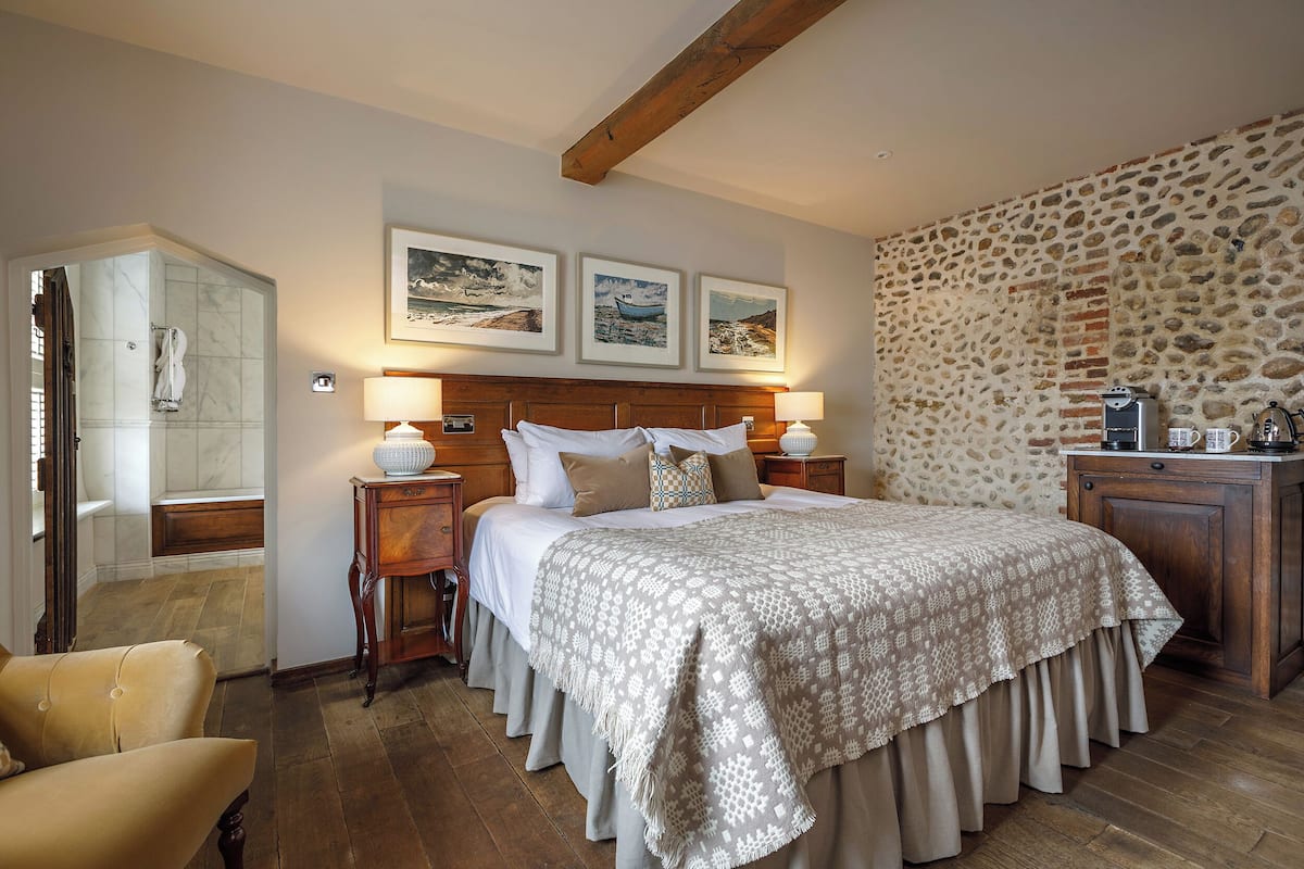 A bedroom with a large bed, wooden headboard, and a stone wall. There are framed pictures on the wall, a nightstand, and a chair.