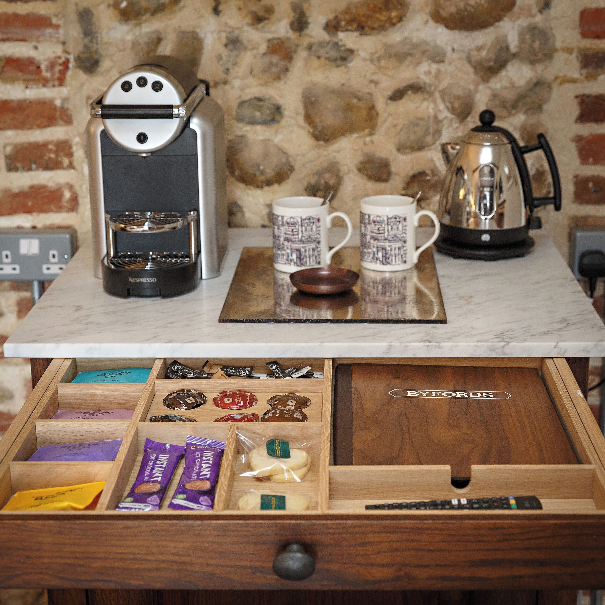 A kitchen counter with a coffee maker, two mugs, and a kettle. Below, a drawer contains various food items and a remote control.