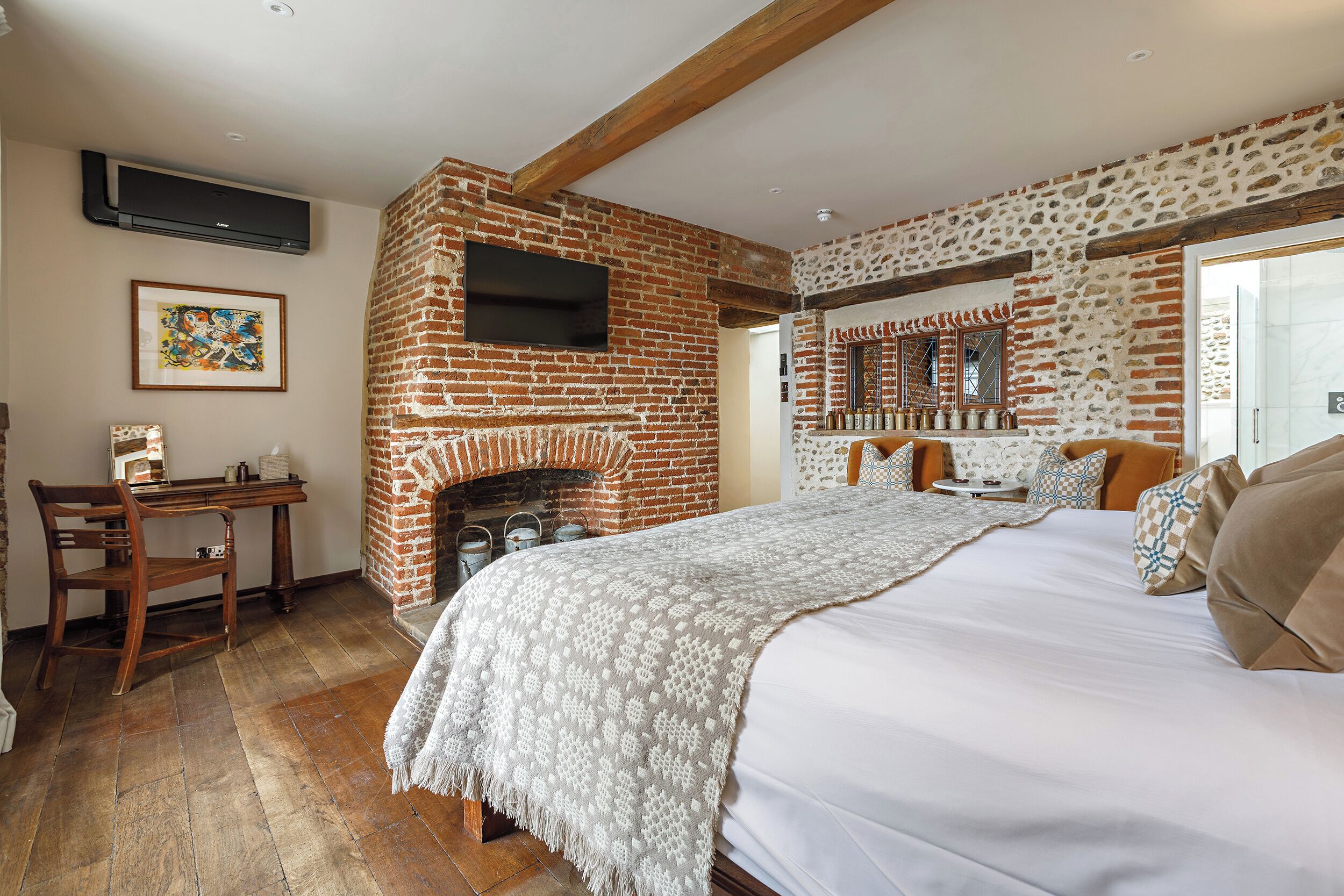 Un dormitorio con chimenea de ladrillo, una cama con manta estampada, un escritorio con silla y un televisor montado en la pared.