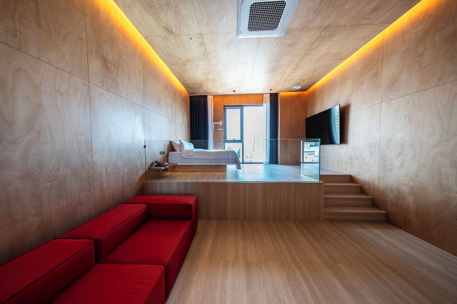 A modern hotel lobby with a wooden floor, a red sofa, a bed with a city view, and a TV mounted on the wall.