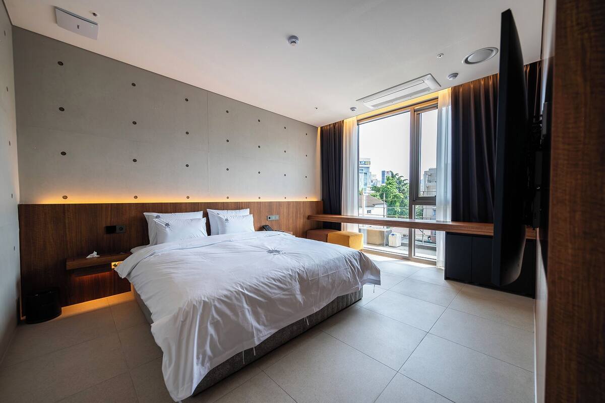 Una habitación de hotel moderna con una cama grande, cabecera de madera, mesita de noche y cortinas en la ventana, con vistas a la ciudad.