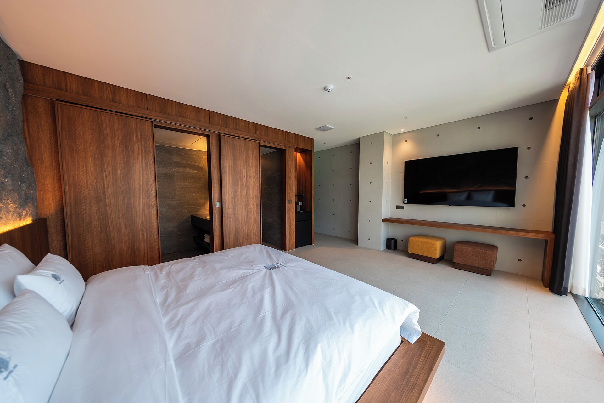 A modern hotel room with a large bed, wooden wardrobe, a flat-screen TV mounted on the wall, and a small seating area.