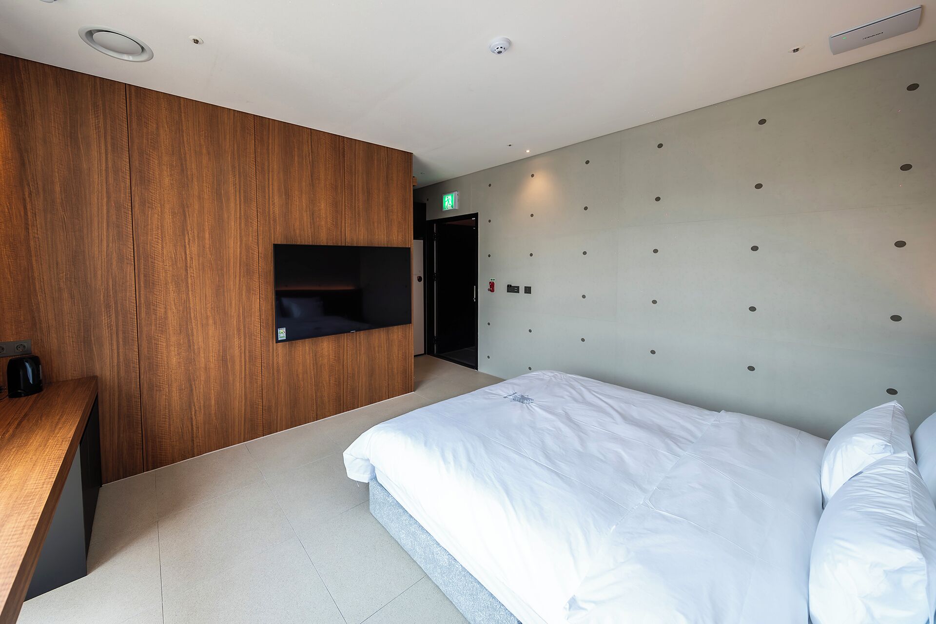 Una habitación de hotel moderna con una pared de acento de madera, una estantería empotrada, una cama con ropa de cama blanca y una lámpara de pared.