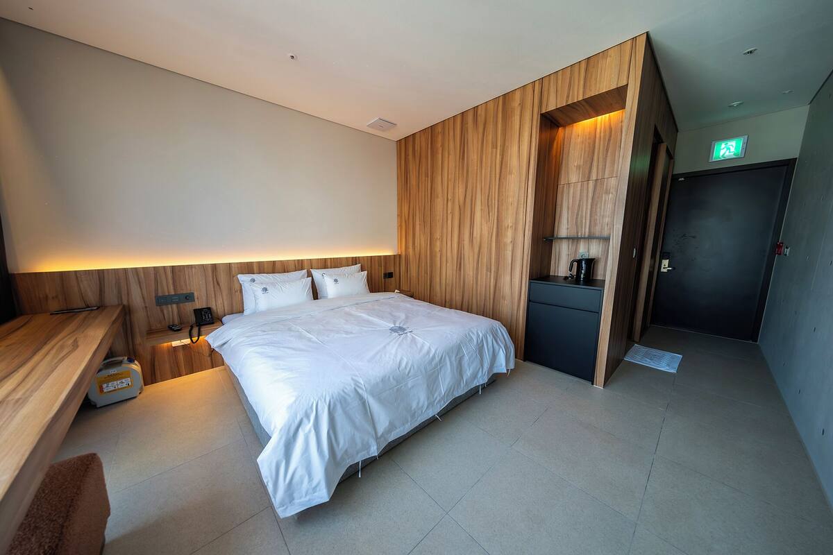 Una habitación de hotel moderna con una cama grande, paneles de madera, un escritorio pequeño y la puerta del baño.