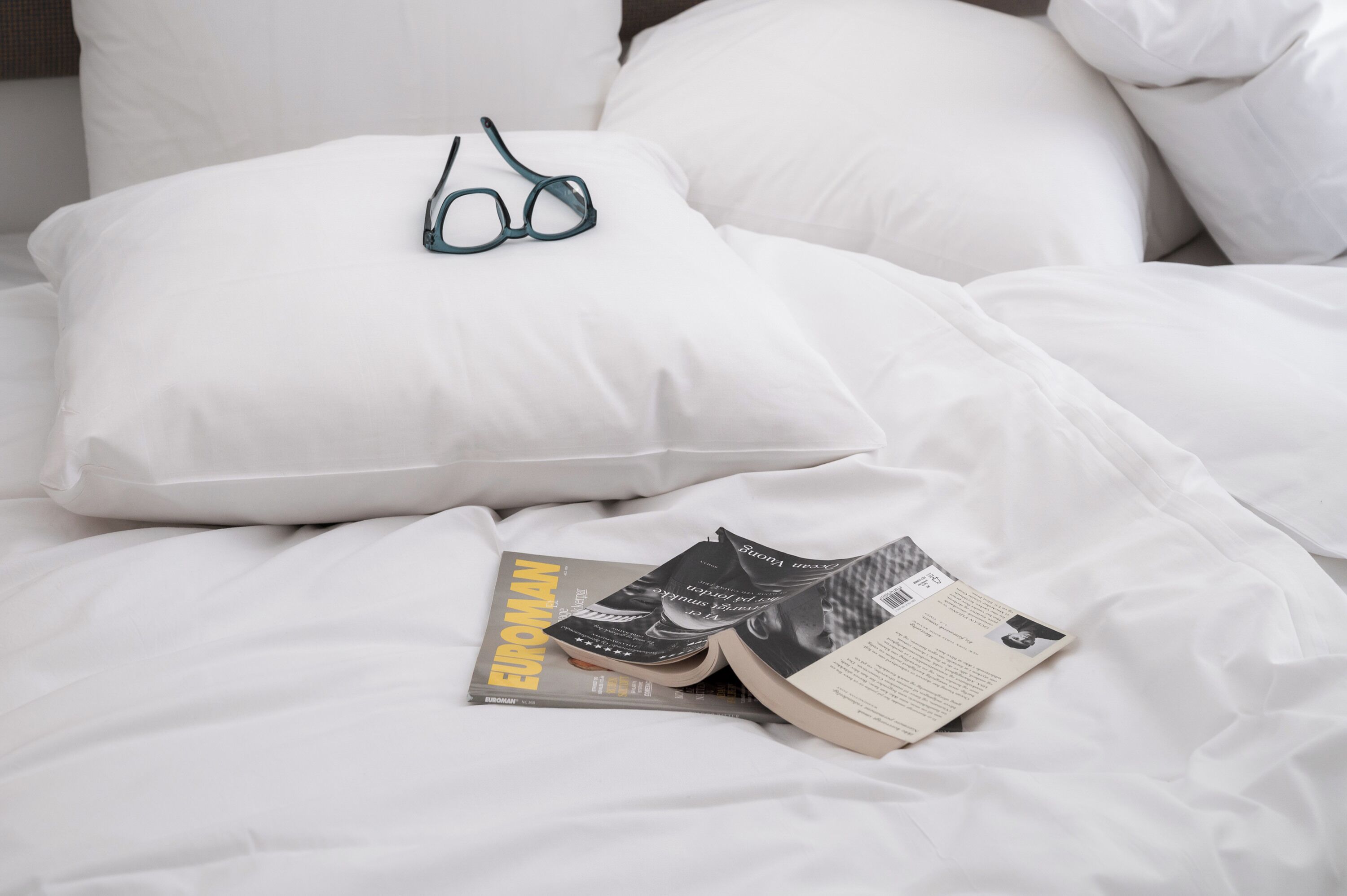 Una cama con ropa de cama blanca, un par de gafas y una revista titulada 'EUROMAN'.