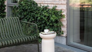 Un bâtiment en briques avec une fenêtre, un banc vert et un piédestal blanc sur lequel sont posés une tasse et une soucoupe.