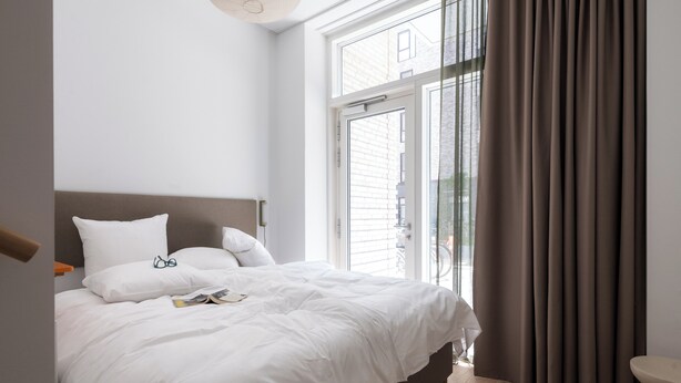 Une chambre à coucher moderne avec un grand lit, du linge de lit blanc, une table de chevet et une fenêtre avec des rideaux.