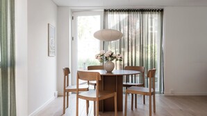 Un coin repas comprenant une table ronde, quatre chaises en bois, un vase avec des fleurs et une grande fenêtre avec des rideaux.
