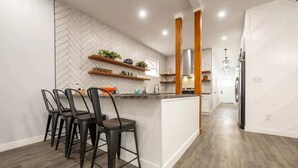 Eine moderne Küche mit einem Zickzack-Muster an der Spritzschutzwand, Holzregalen mit Pflanzen, einer zentralen Kochinsel inklusive Barhocker und einem Edelstahl-Kühlschrank.