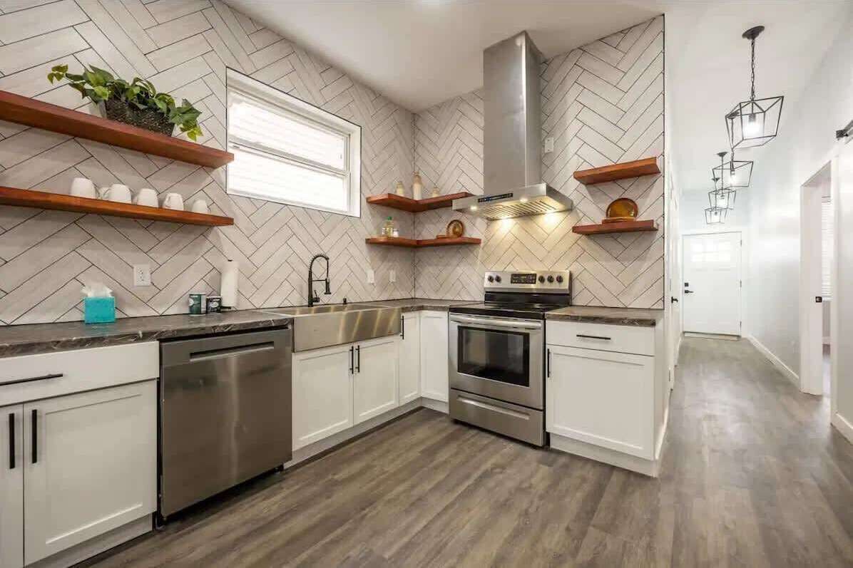 Una cucina moderna con mobili bianchi, lavastoviglie in acciaio inox, forno integrato e zoccolo a mosaico a spina di pesce.
