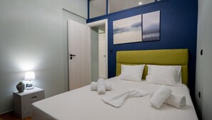 Un lit bien fait, avec du linge de lit et des serviettes blanches, une table de chevet avec une lampe, et une porte donnant sur une autre pièce.