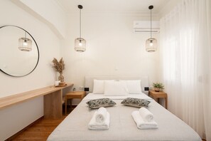 A hotel room with a bed, two bedside tables, a round mirror, a wall-mounted air conditioner, and two hanging pendant lights. - LIV - Charming 3BD in Athens Vibrant Downtown (Athens)