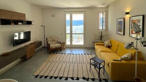 A modern living room with a flat-screen TV, a yellow sofa, a patterned rug, and a view of the sea through a glass door.
