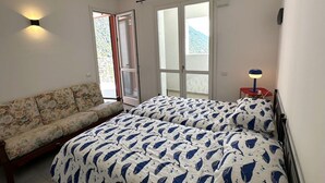 A bedroom with a sofa, two beds with matching bedding, a bedside table with a lamp, and a window with blinds.