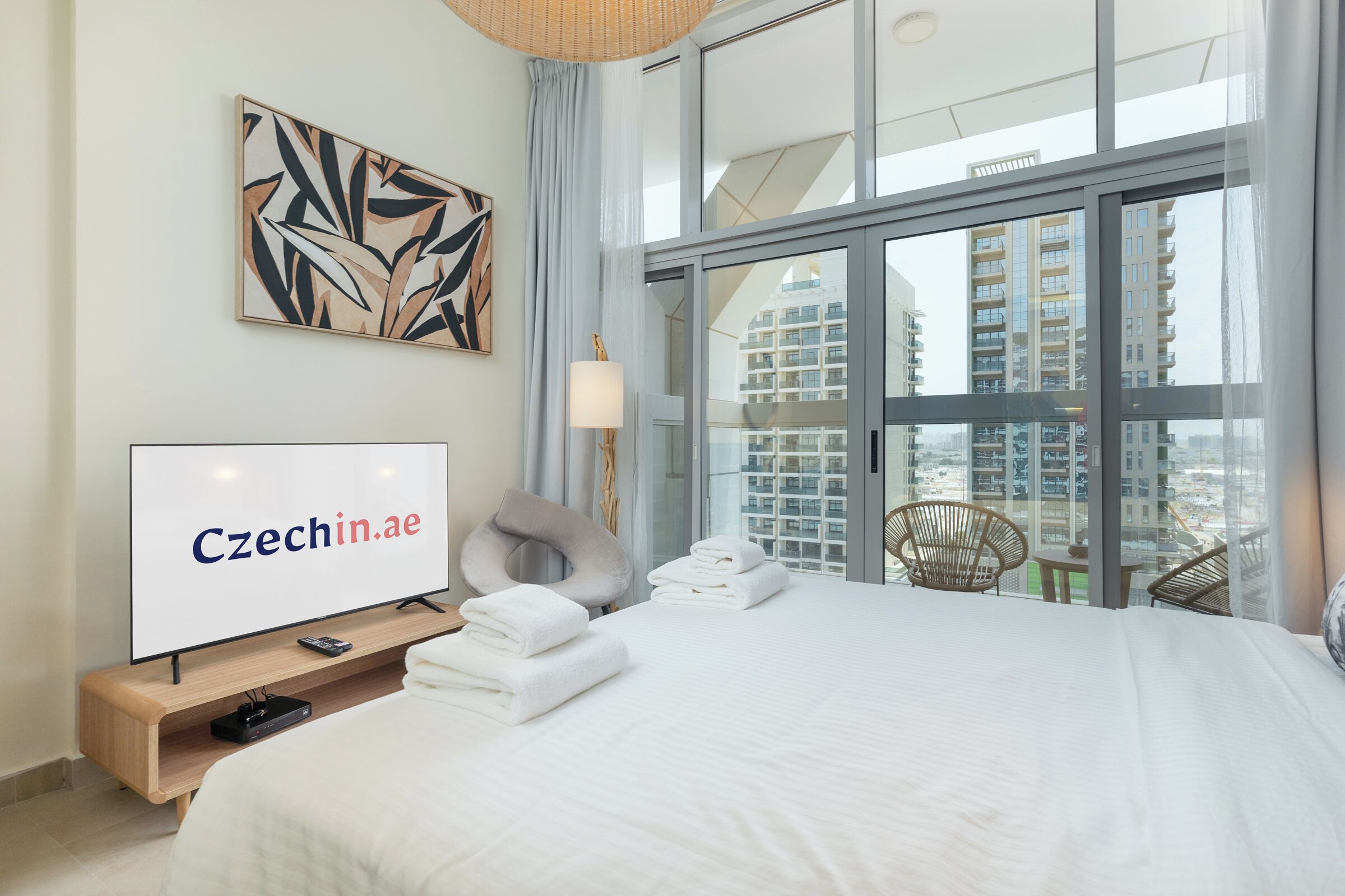 A modern hotel room with a large bed, a flat-screen TV, a view of high-rise buildings, and a balcony with wicker furniture.