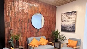 Ein moderner Empfangsbereich mit Holzbalkendecke, einem runden Spiegel und einem Bild an der Wand. Zwei graue Sofas mit orangefarbenen Kissen, ein kleiner Tisch und Topfpflanzen sind ebenfalls vorhanden.