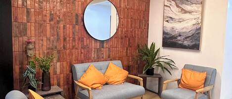 Un área de recepción moderna con techo de madera, un espejo redondo y un cuadro en la pared. Hay dos sofás grises con cojines anaranjados, una mesita y plantas en macetas.