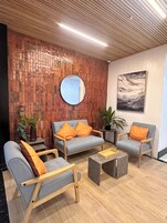 Un área de recepción moderna con techo de madera, un espejo redondo y un cuadro en la pared. Hay dos sofás grises con cojines anaranjados, una mesita y plantas en macetas.