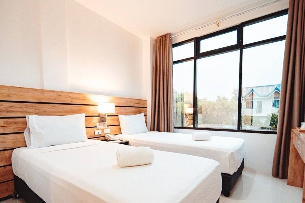 Une chambre d’hôtel avec deux lits, une tête de lit en bois, une grande fenêtre avec des rideaux et une vue sur les bâtiments environnants.