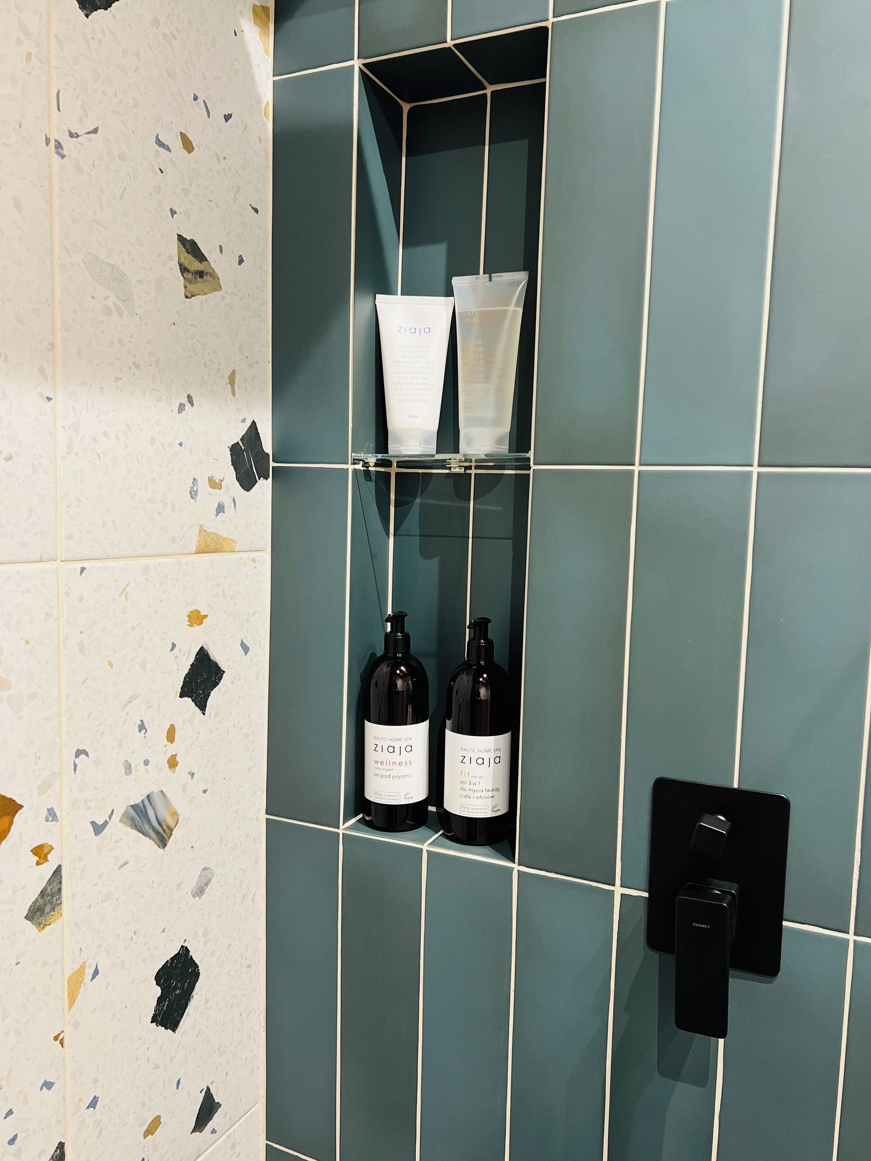 Un baño con pared de azulejos, estante con botellas y un interruptor de luz.