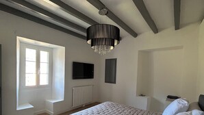 A bedroom with a large bed, a chandelier, a window, a radiator, and a television on the wall. - Village house in the heart of Castagniccia (CARCHETO)