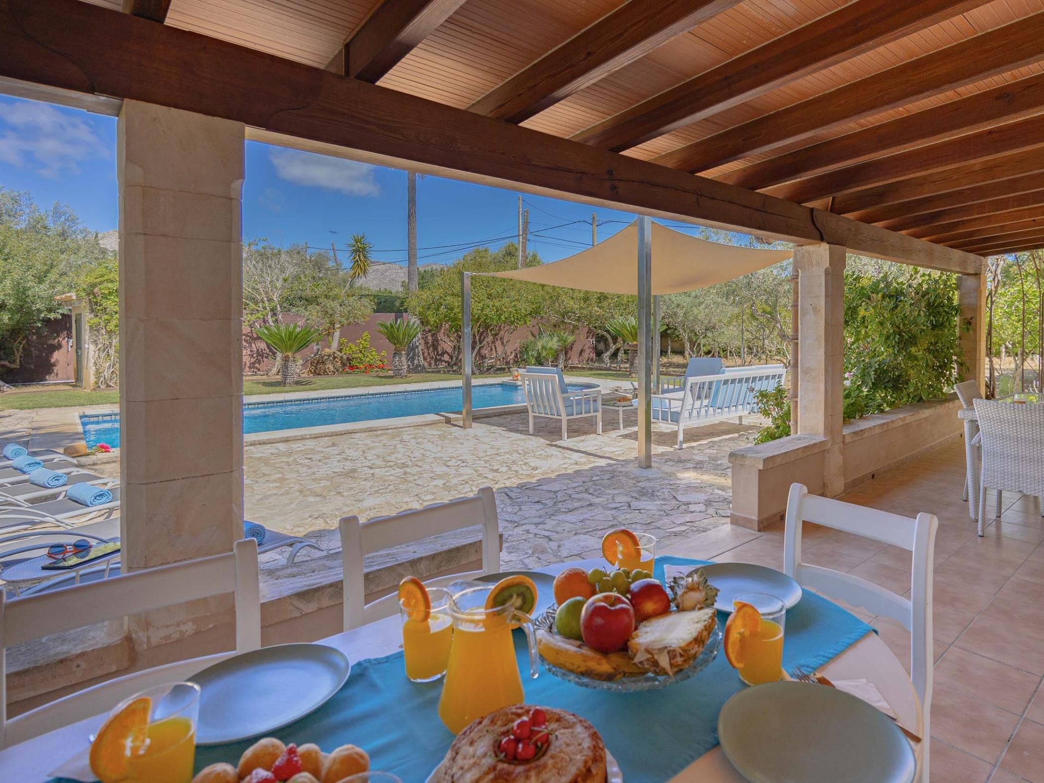 Un espace extérieur abrité, aménagé pour les repas, avec vue sur la piscine, un sol en pierre et une table dressée avec des fruits et des boissons.