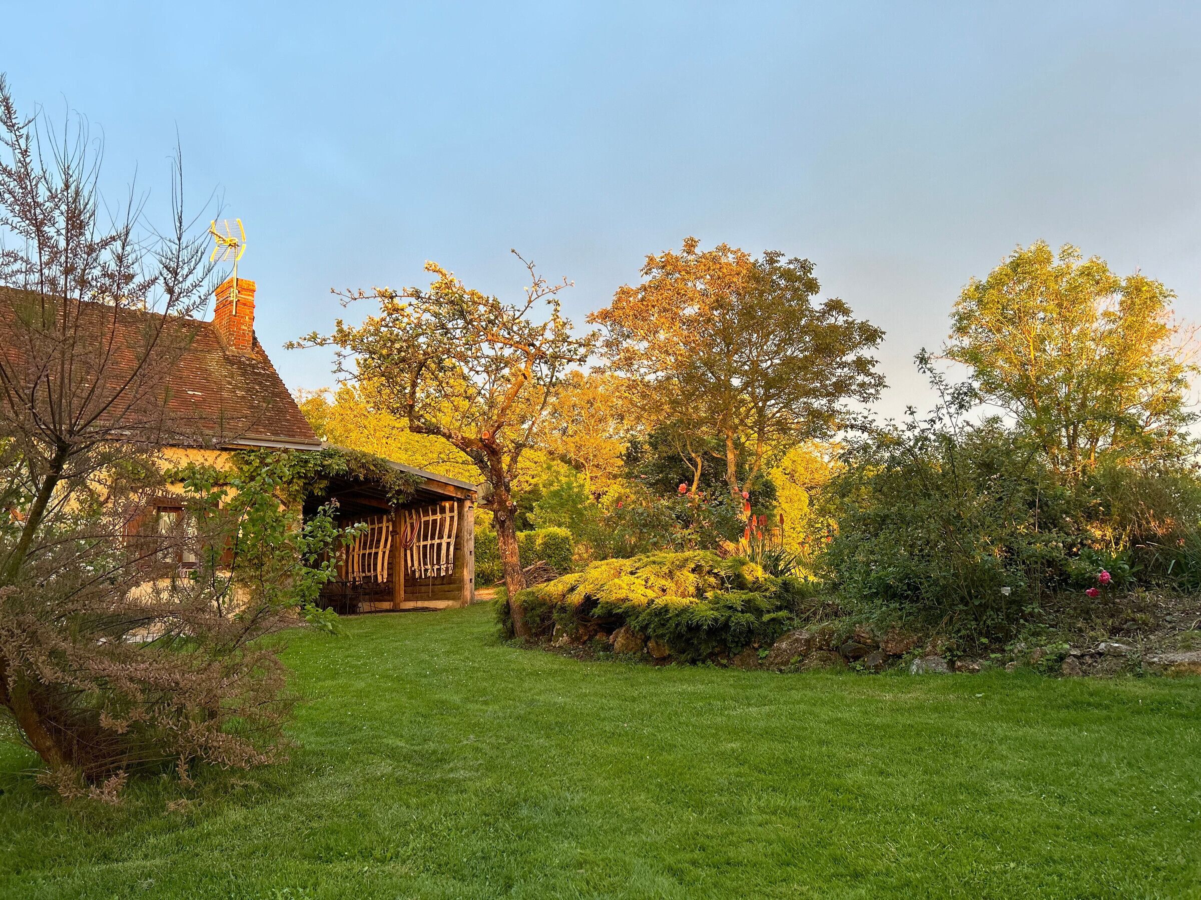 A rustic house with a sloped roof and a chimney, surrounded by a well-maintained garden with various shrubs and flowering plants.