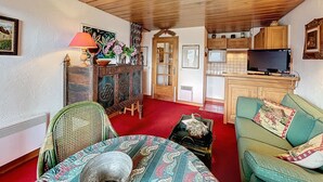 A cozy living room with a wooden ceiling, a dining area with a patterned tablecloth, a green sofa with decorative pillows, and a kitchen area with wooden cabinets and a flat-screen TV. - 2-room apartment with parking, near the slopes (Combloux)