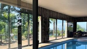 Ein moderner, überdachter Pool mit Glaswänden, einer Holzterrasse und Blick auf üppige Grünflächen.