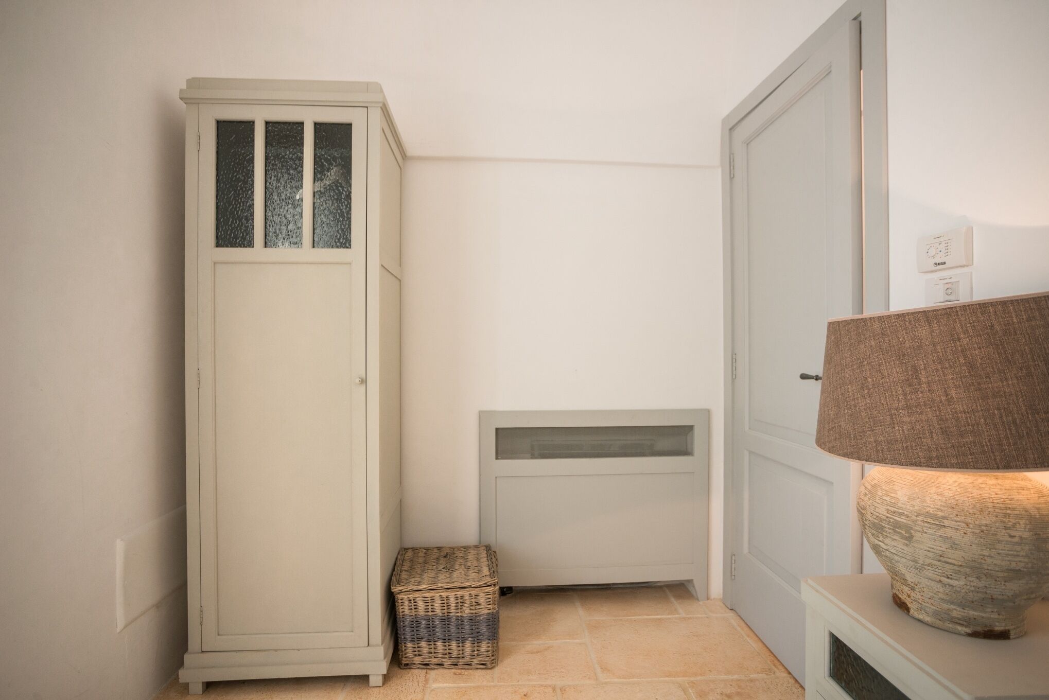Habitación de esquina con armario blanco, una pequeña cesta de mimbre, un radiador y una lámpara sobre una mesita de noche.