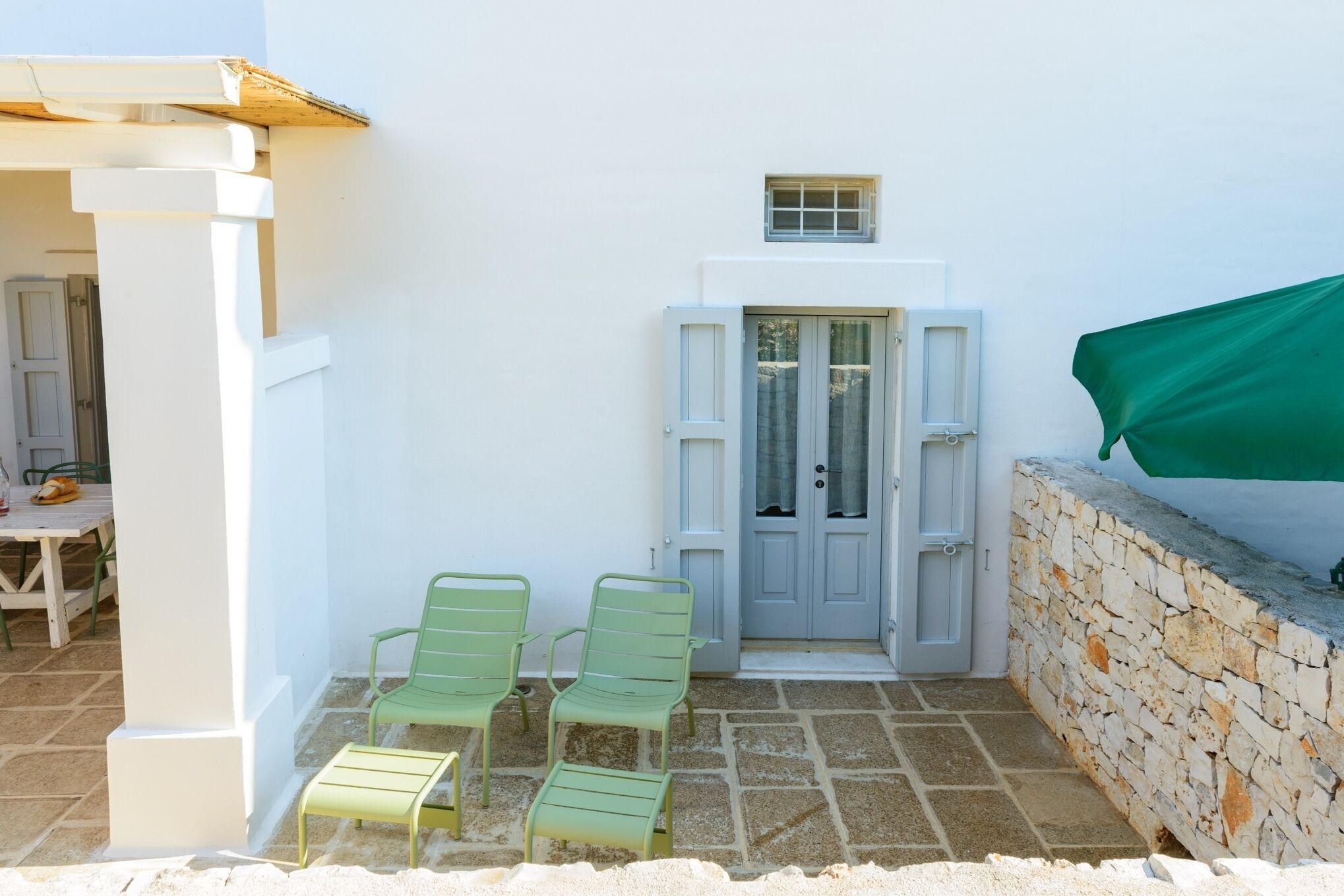 A white building with a stone wall, a door with shutters, and two green chairs on a tiled patio.
