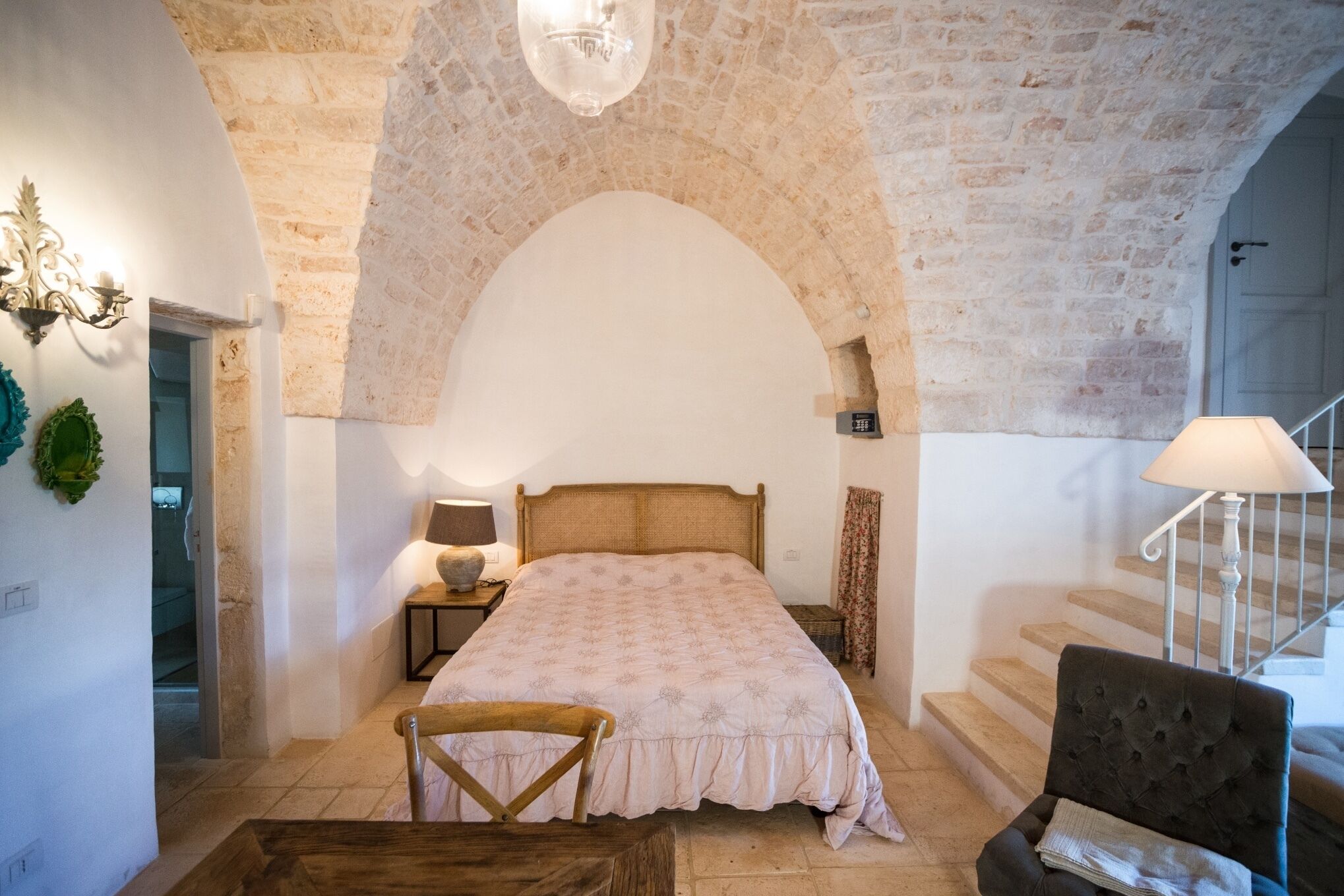 Un dormitorio con un gran techo de piedra en arco, una cama con colcha estampada, una silla de madera, una mesita de noche con lámpara y una escalera con barandilla blanca.