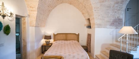 Un dormitorio con un gran techo de piedra en arco, una cama con colcha estampada, una silla de madera, una mesita de noche con lámpara y una escalera con barandilla blanca.
