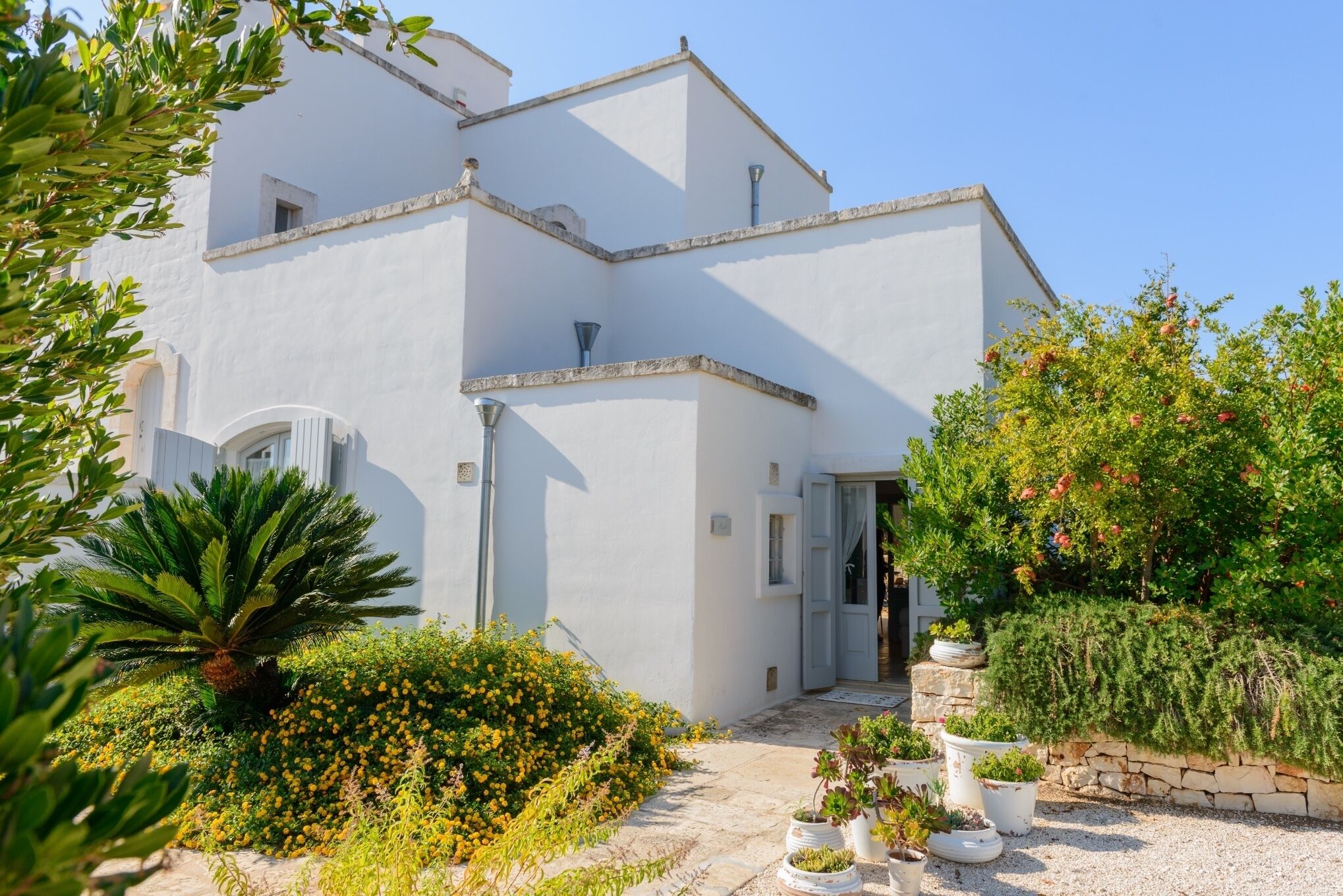 A white building with a stone facade, a small garden with various plants, and a clear sky.