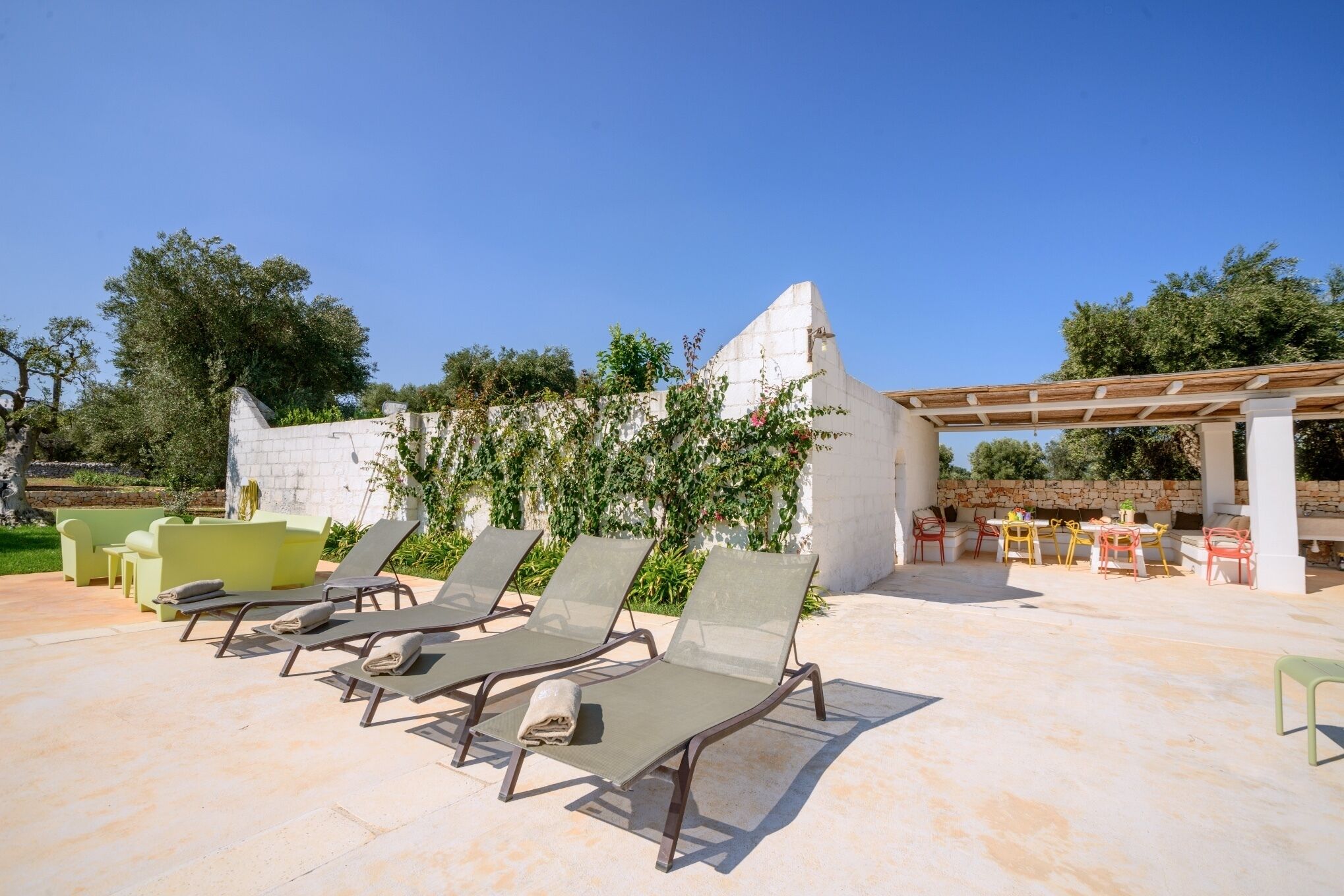 A sunlit outdoor seating area with lounge chairs and tables, surrounded by greenery and a stone wall.