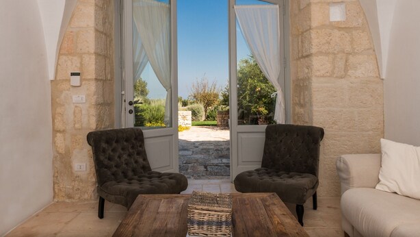 A cozy room with a stone wall, two dark armchairs, a wooden table, and a view through a glass door to an outdoor garden. - Masseria Pepenofio (Monopoli)