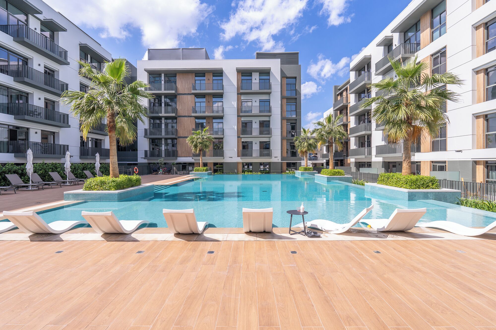 A modern apartment building with a central swimming pool, lounge chairs, and palm trees.