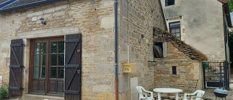 A rustic stone building with a wooden door and shutter, a small outdoor seating area with plastic chairs and a table, and a barbecue grill.