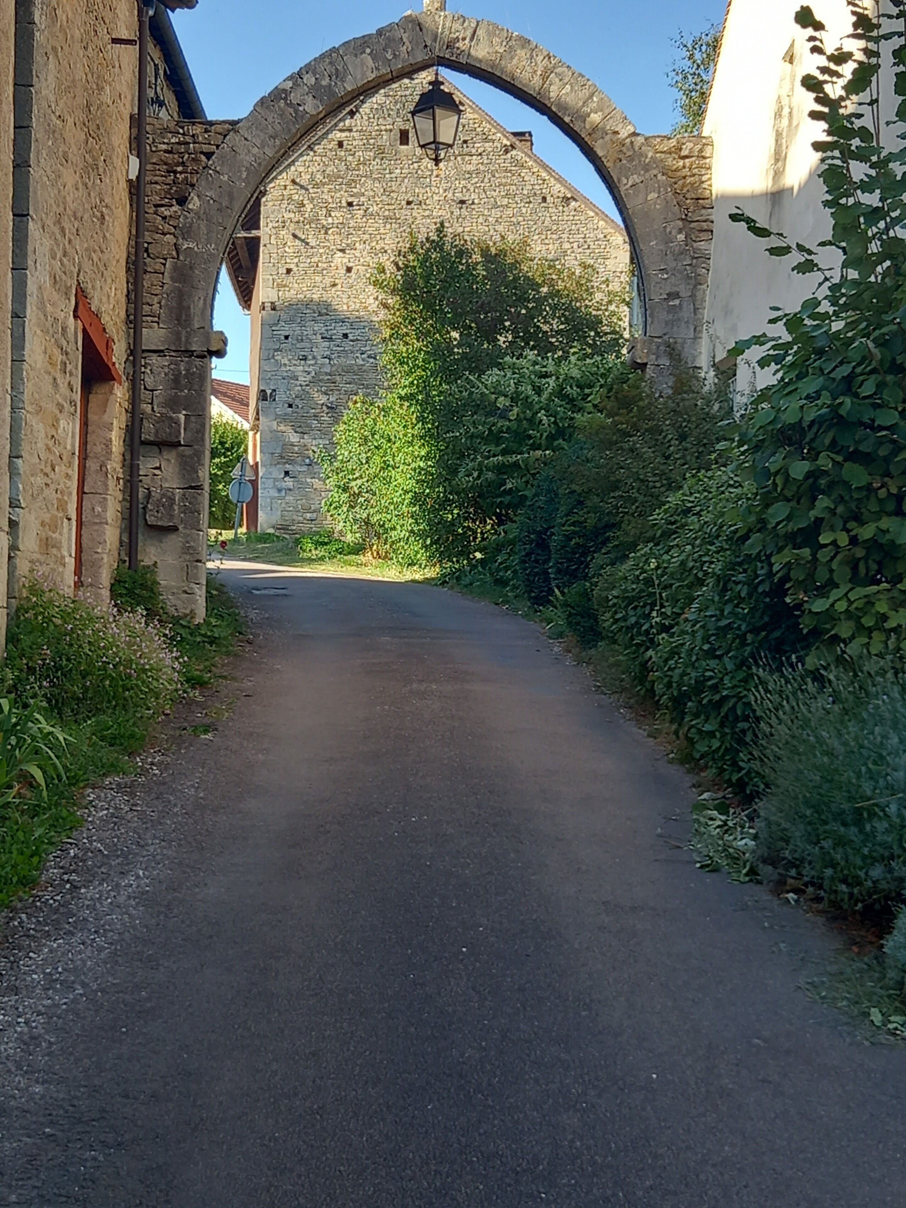 Un camino pavimentado y estrecho bordeado por edificios de piedra y vegetación, que conduce a un arco de piedra con un farol.