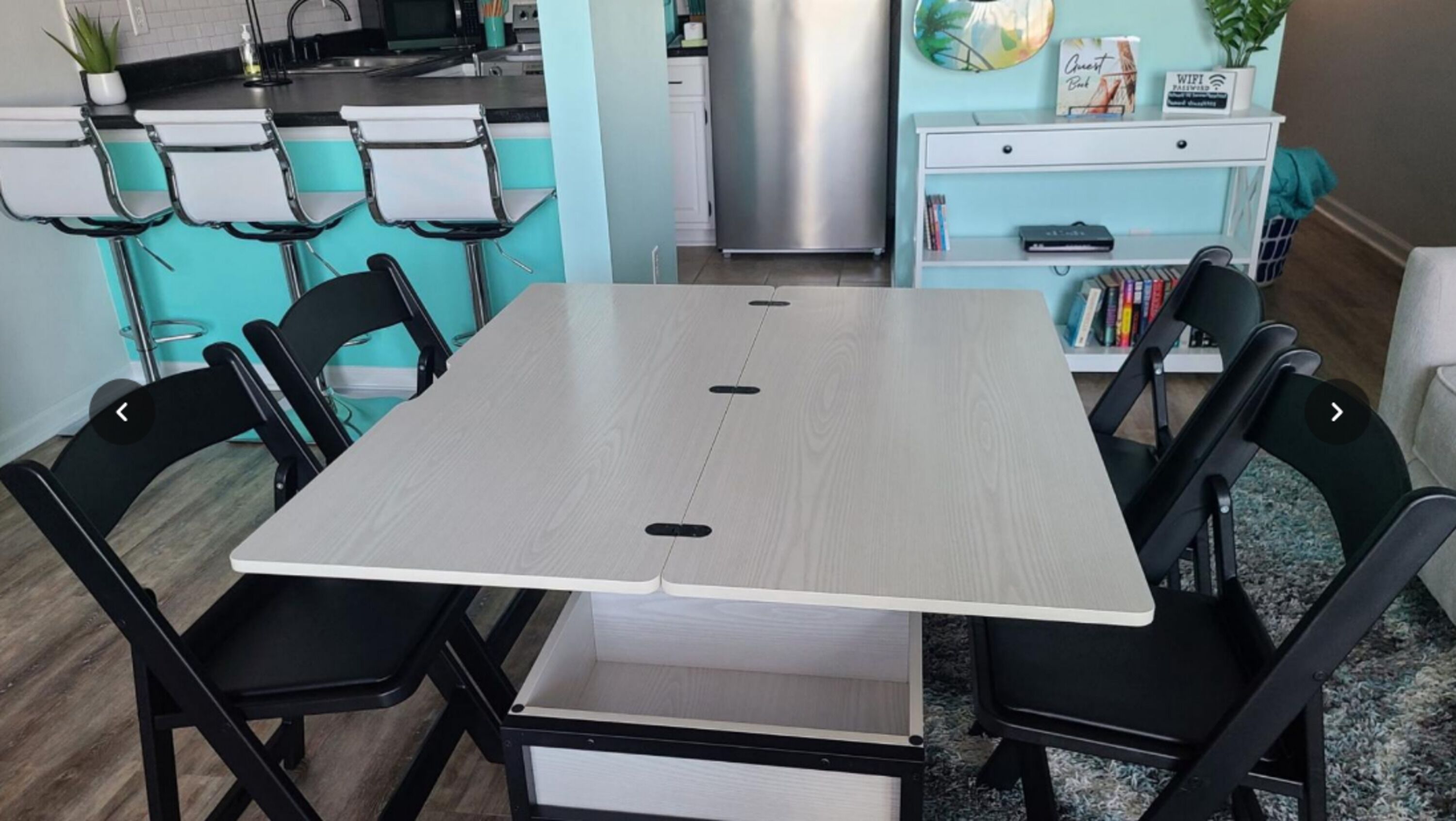 A dining area with a light wood table, black chairs, a white cabinet, and a kitchen in the background.