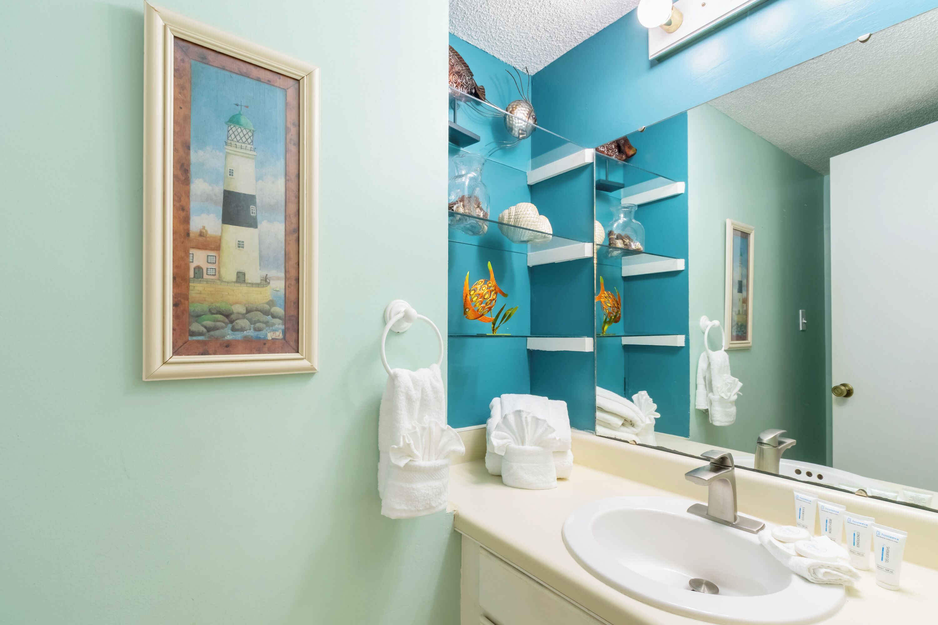 Un baño con un lavamanos blanco, un espejo y un cuadro con una pintura de un faro.